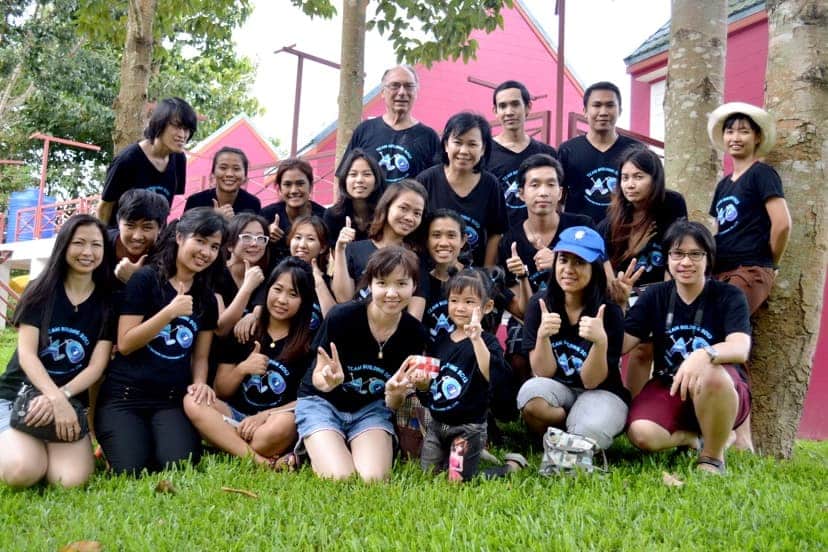A diverse group of people, including men, women, and children, are gathered outdoors, smiling and giving thumbs up. Wearing matching black shirts with a blue design that reflects their shared careers, they sit or crouch on grass with pink buildings and trees behind them.