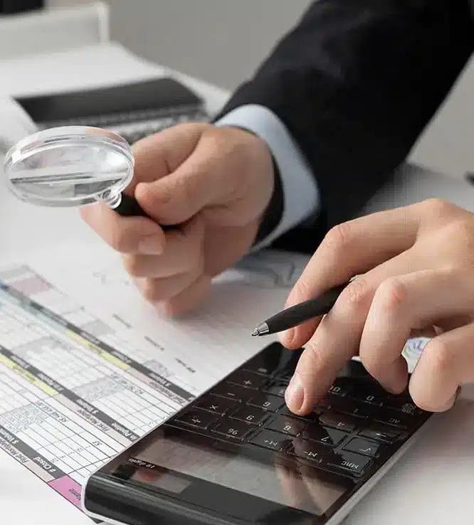 Close-up of a person's hands using a calculator and holding a magnifying glass while reviewing a financial document. The individual, dressed in a suit, suggests they are engaged in professional tax services or financial analysis.