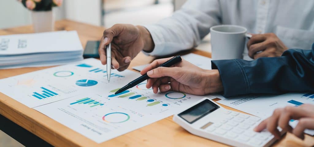 Two people are seated at a wooden table, collaboratively analyzing printed financial charts and graphs. One person is holding a pen and gesturing towards a paper with colorful charts, while the other holds a calculator. A coffee mug and documents are also on the table.