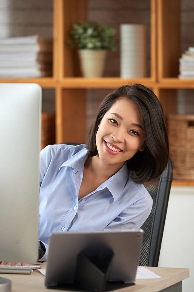 A woman with shoulder-length dark hair, wearing a light blue button-up shirt, is smiling while sitting at a desk with a computer and a tablet. Perhaps contemplating new career opportunities, she’s surrounded by wooden shelves filled with books and a plant in the background.