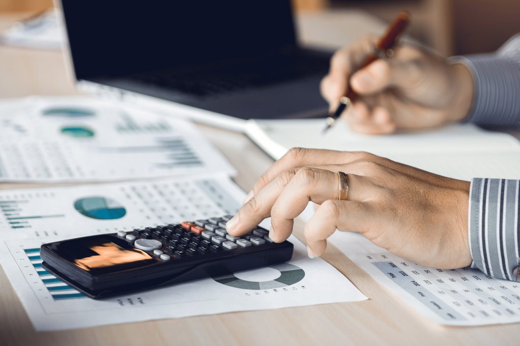 A person wearing a striped shirt is using a calculator on a desk filled with financial documents and charts. One hand is on the calculator, and the other is holding a pen, taking notes on a notepad.