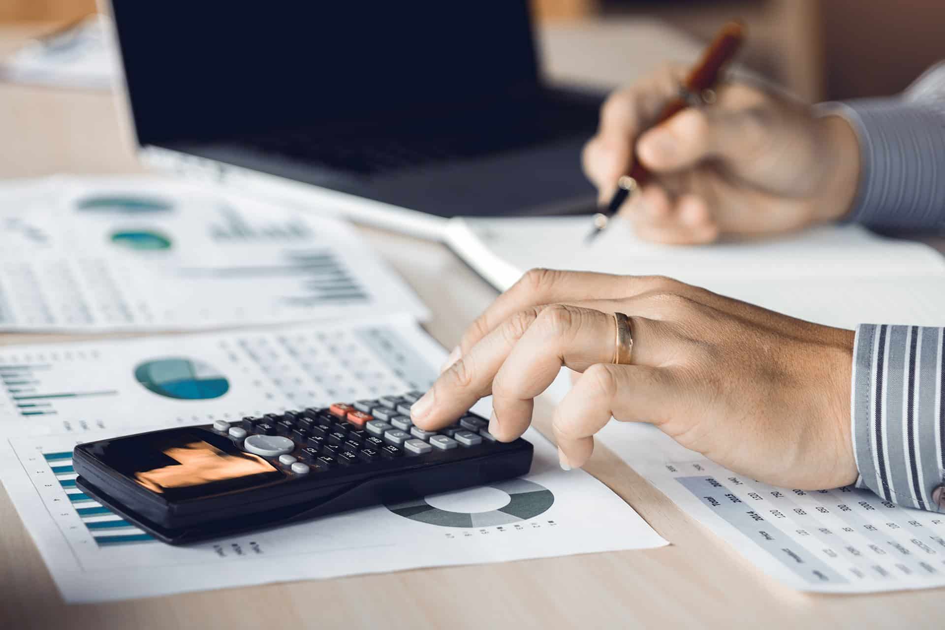 A person wearing a striped shirt is using a calculator on a desk filled with financial documents and charts. One hand is on the calculator, and the other is holding a pen, taking notes on a notepad.