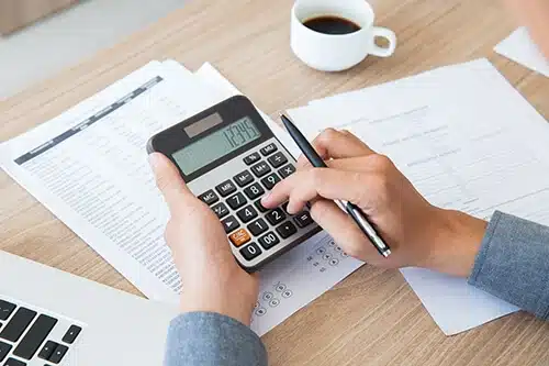 Payroll Outsourcing Service Provider A person is using a calculator on a desk with paperwork, a pen, and a cup of coffee. A laptop is partially visible on the left side of the desk. The scene depicts someone working on financial or business calculations, possibly utilizing outsourced accounting services.