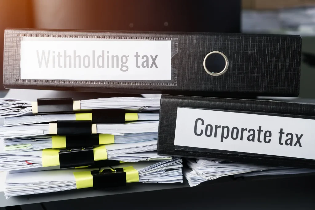 A stack of neatly organized documents secured with binder clips sits on a desk. Two large black binders labeled "Income Tax in Thailand" and "Corporate tax" are positioned on top of the stack, with natural light illuminating the scene from the left.