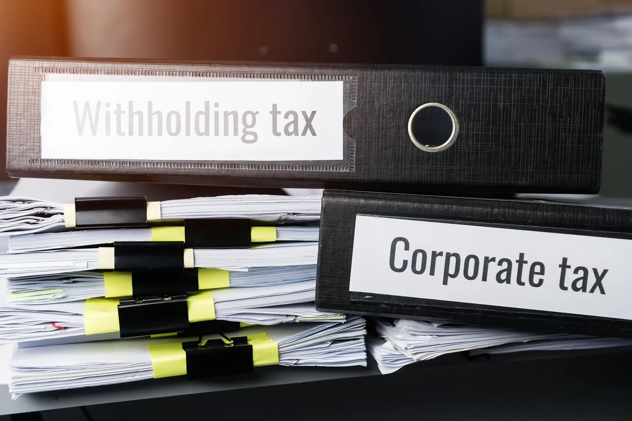 A stack of neatly organized documents secured with binder clips sits on a desk. Two large black binders labeled "Income Tax in Thailand" and "Corporate tax" are positioned on top of the stack, with natural light illuminating the scene from the left.