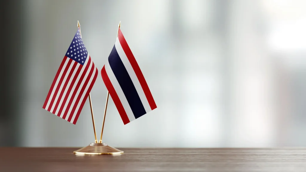 Two small flags are crossed and displayed on a wooden surface with a blurred background. The flag on the left is the United States flag, while the flag on the right is the Thai flag. This display symbolizes the strong ties established through the USA Treaty of Amity, fostering cooperation and friendship.