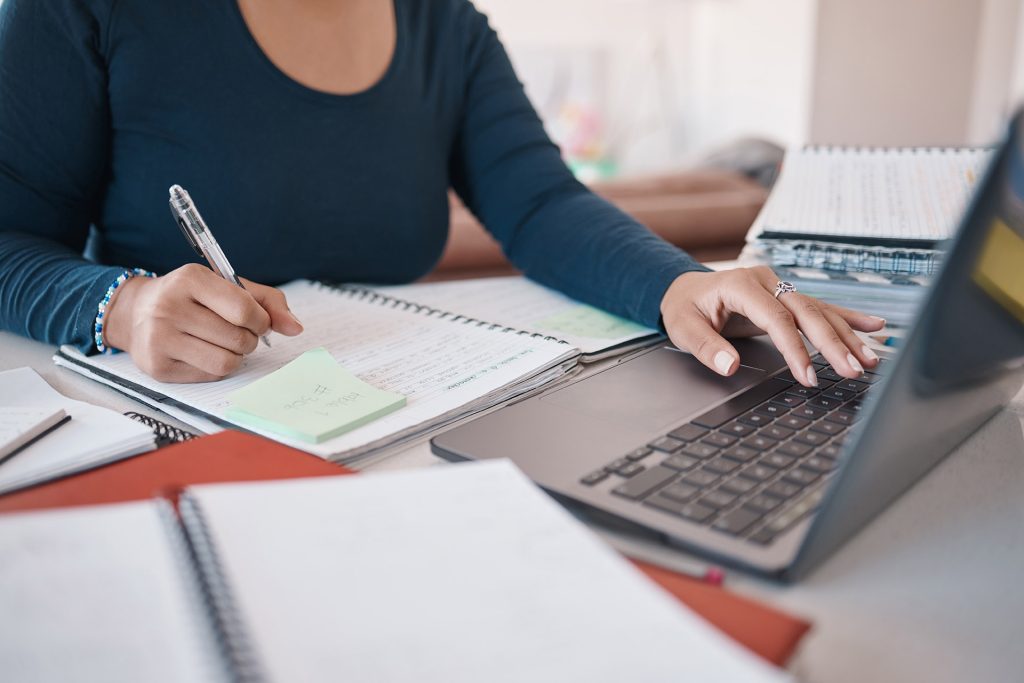 A person with a dark long-sleeve shirt is writing in a notebook with a pen in their right hand while using a laptop with their left hand. Various books and papers are spread across the table.