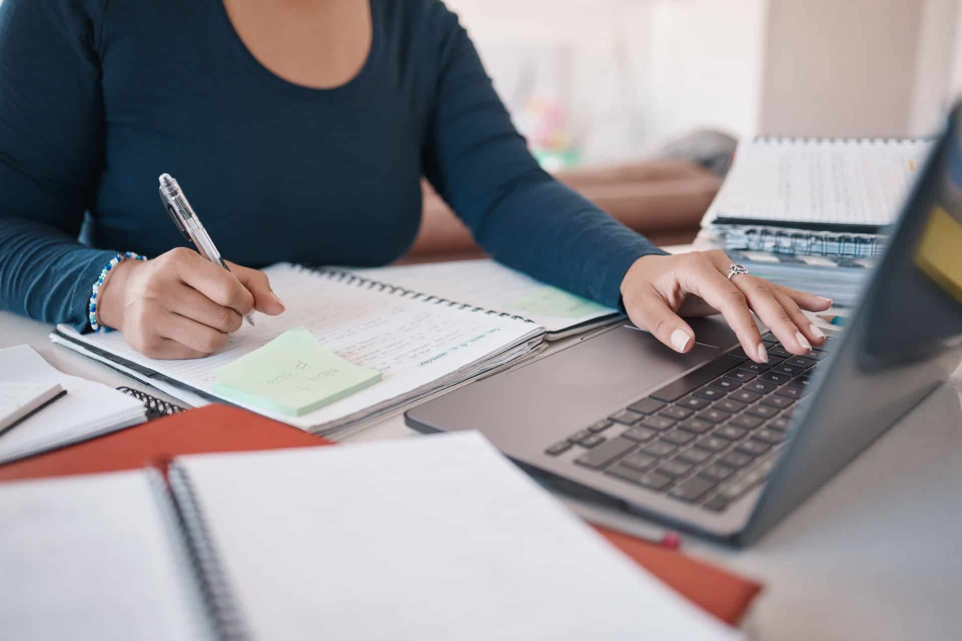 A person with a dark long-sleeve shirt is writing in a notebook with a pen in their right hand while using a laptop with their left hand. Various books and papers are spread across the table.