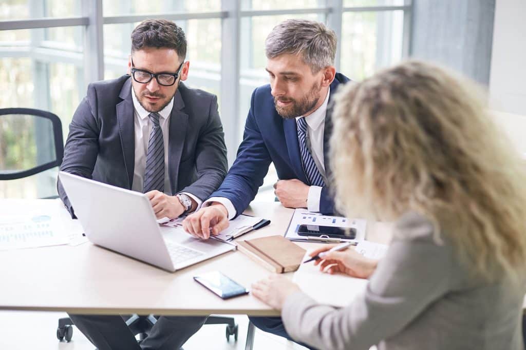 Three business professionals are at a table in a modern office setting. Two men in suits are discussing something shown on a laptop screen, while a woman with curly blonde hair is writing in a notebook. Papers, a smartphone, and a pen are also on the table.