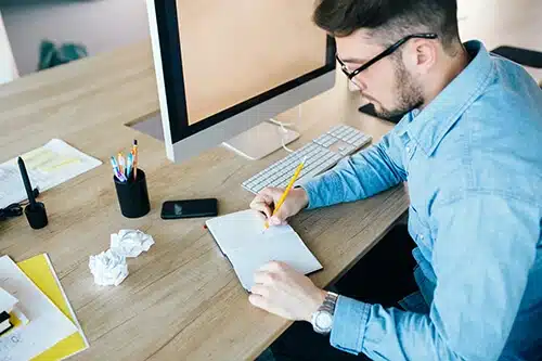 A man wearing glasses and a blue shirt sits at a wooden desk, writing in a notebook with a yellow pencil. A desktop computer displaying accounting services, smartphone, cup of colorful pens, and crumpled papers are also on the desk.