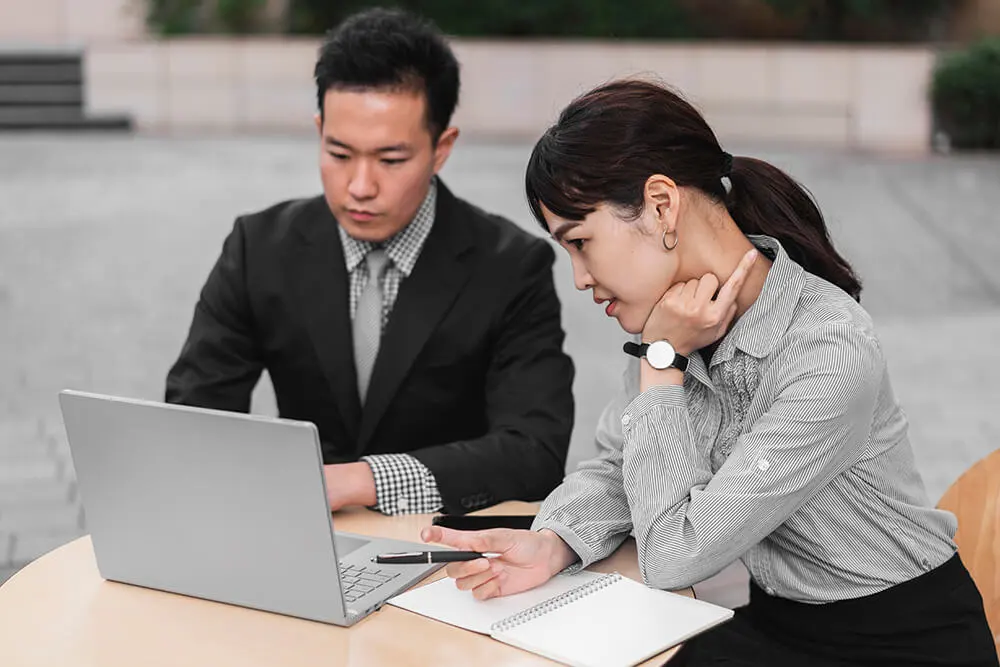 Two individuals are sitting at a table outdoors and looking at a laptop. The man is wearing a suit and tie, while the woman, who has a ponytail and is holding a pen, is wearing a striped shirt and has a notebook in front of her. They appear to be focused on discussing outsourced accounting and Advisory Ltd.