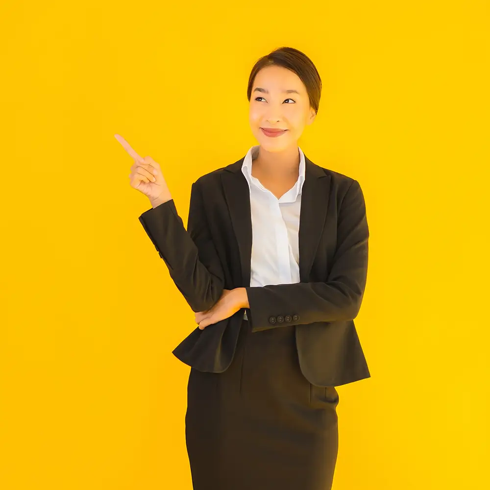 A woman in a black suit and white shirt stands against a bright yellow background, smiling and pointing to her left with her right hand while her left arm is crossed over her waist, embodying the professionalism of top-tier outsourced accounting.