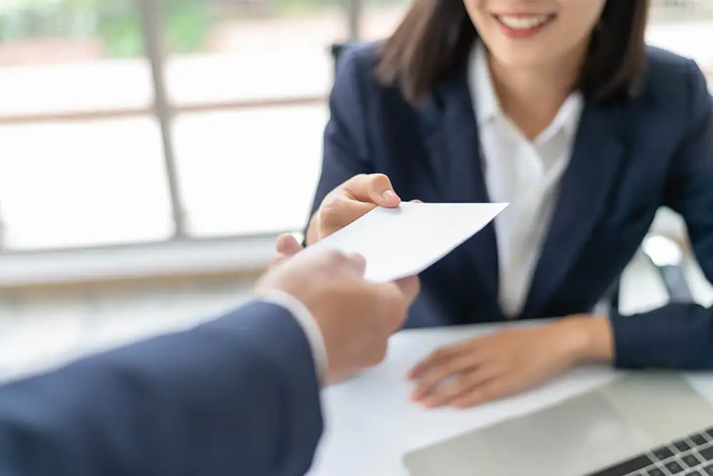 A person in a suit hands an envelope to a smiling woman across a table. She is also dressed in business attire and is seated beside a laptop, likely discussing outsourced accounting services. The setting appears to be a professional office environment.