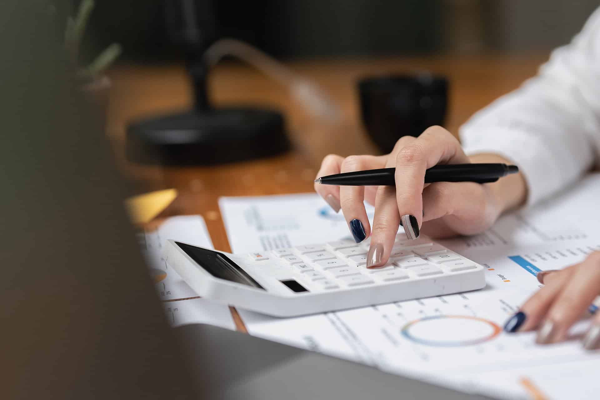 Person using a white calculator while holding a pen. Various documents with charts and graphs are spread out on the desk. Only the person's hands and part of their arms are visible. A coffee mug and a blurred background can also be seen. Strong Growth in Thailand