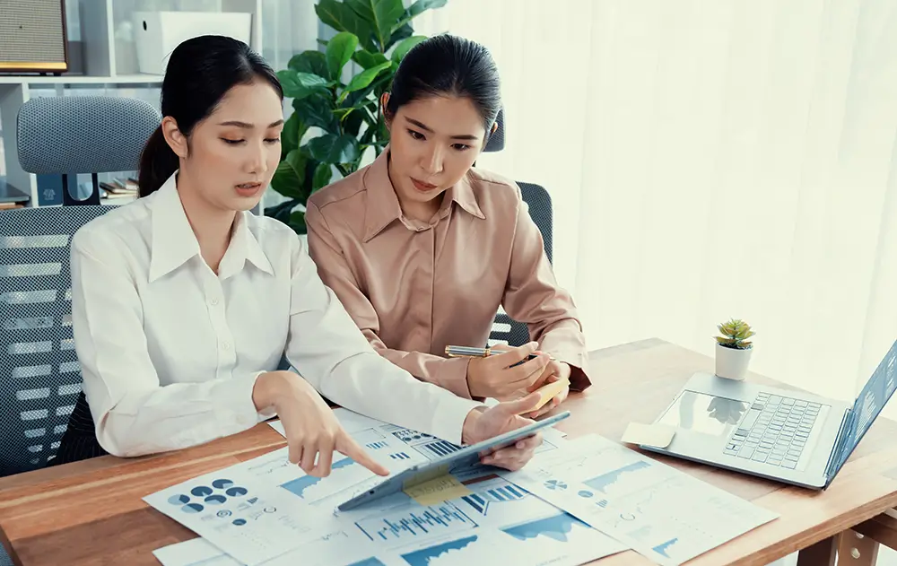 Two women are seated at a wooden desk, viewing and discussing documents filled with charts and graphs. One woman points to a section on corporate tax planning while the other pays attention. A laptop is open on the desk with some office supplies and a small plant nearby.
