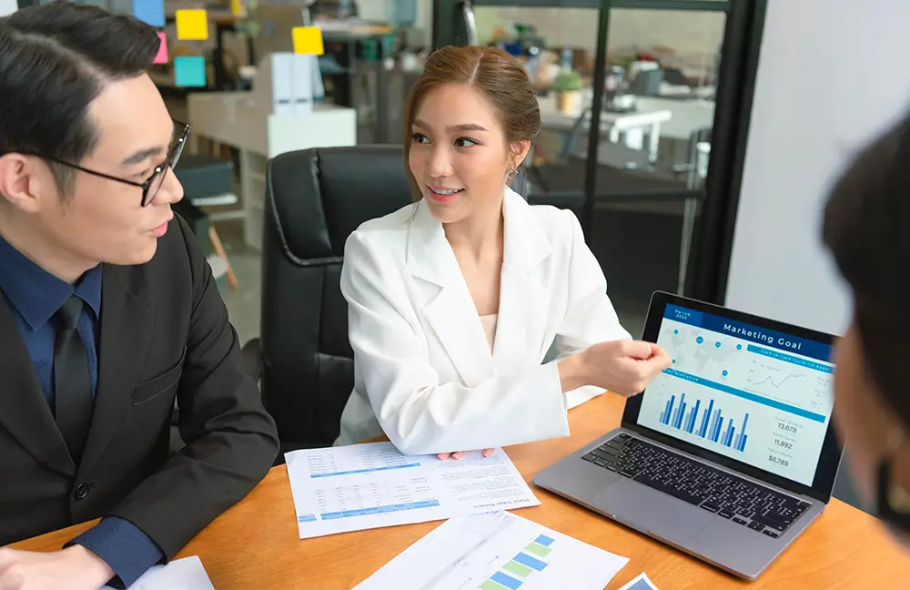 A woman in a white blazer sits at a table, pointing at a laptop screen displaying various charts and graphs labeled 'Marketing Goals.' She is discussing Expert Corporate Accounting Services with a man in a dark suit seated next to her. Papers with charts are spread out on the table. Mergers and Acquisitions