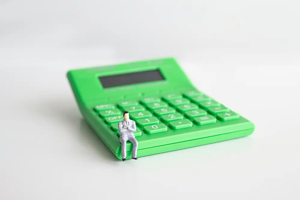 A small figurine of a person in a suit and tie is sitting with crossed arms on the corner of a large green calculator, which dominates the background. The scene playfully underscores the expertise in corporate accounting services, highlighting the tiny figure's insignificance next to the oversized calculator.